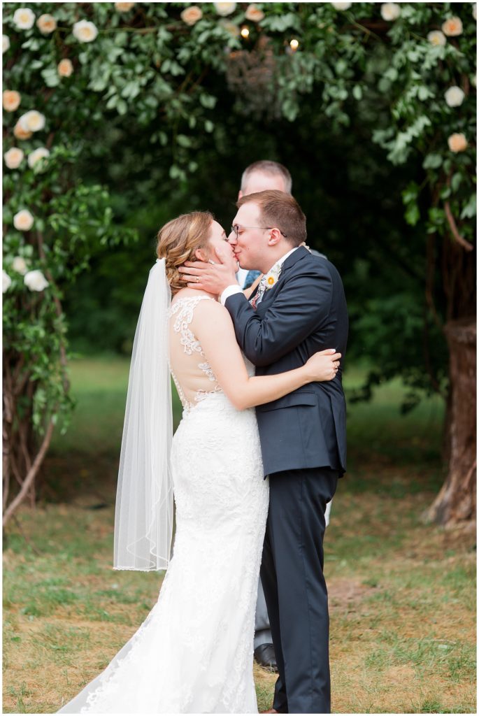 Just married couple kiss at outdoor reception at Hope Glen Farm under floral archway by Essen Events