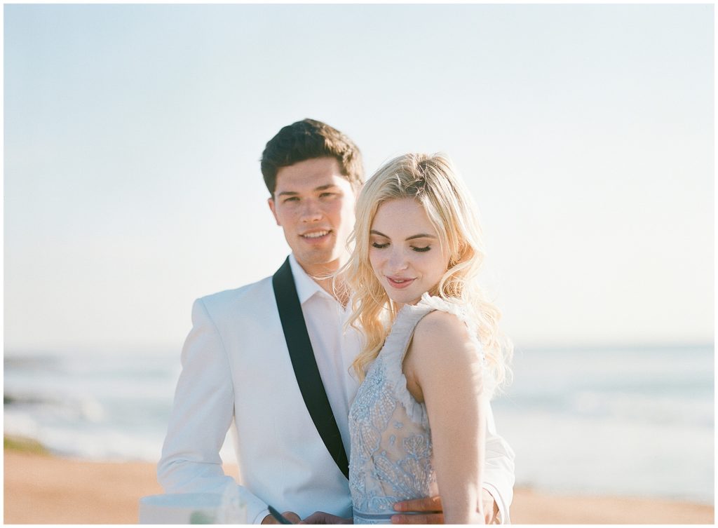 A bride and groom stand in front of an ocean, the groom is looking at the camera in a Friar Tux suit, and the bride is looking down at the beach.