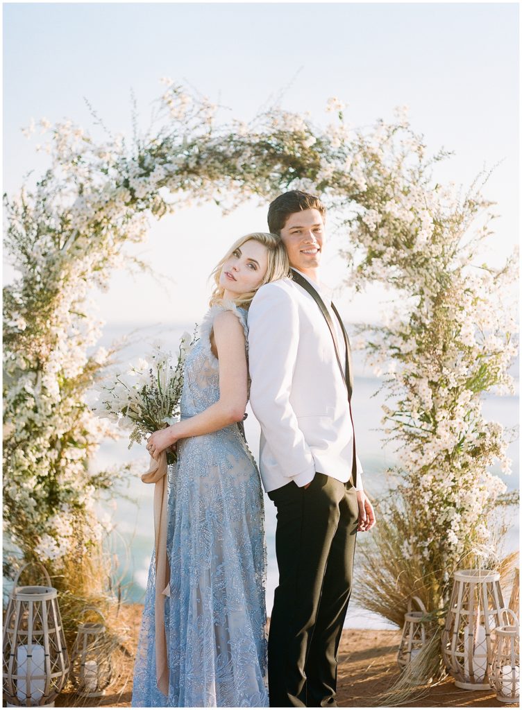 A bride and groom stand back to back under a Max Owens Design white and green archway in front of the Pacific Ocean at sunset