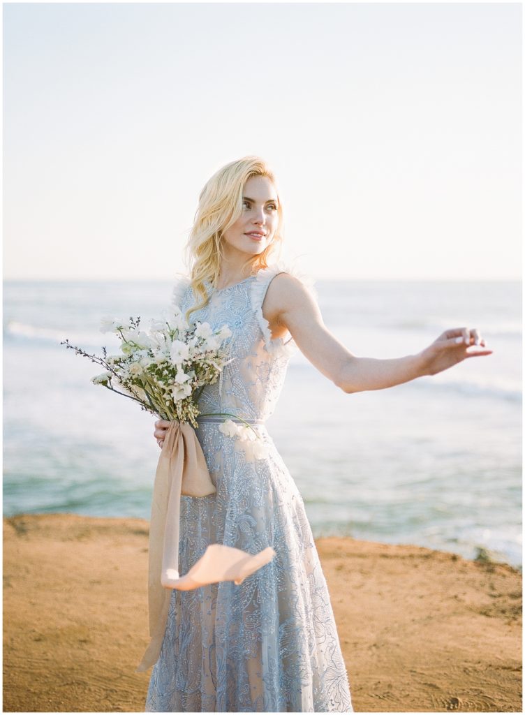 A bride smiles and looks off in the distance holding a gorgeous white floral bouquet with light, sand colored ribbon, which is complimented by a sand beach and light ocean in the background.
