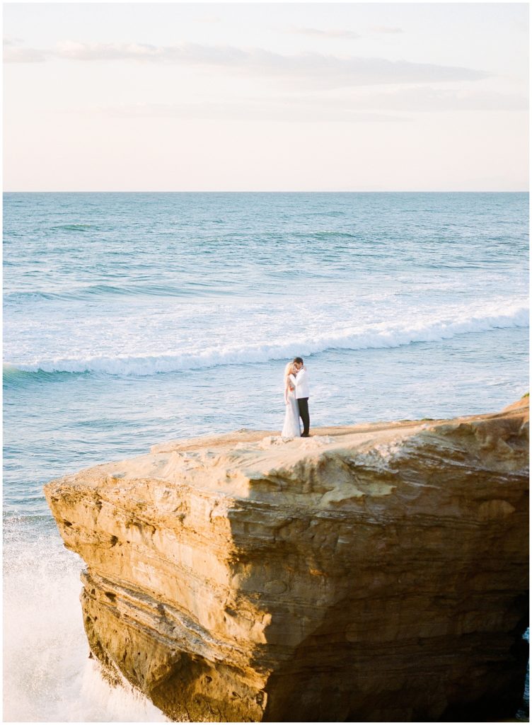A fine art wedding photograph of a couple in the distance, standing on a bluff in front of a warm, blue ocean.