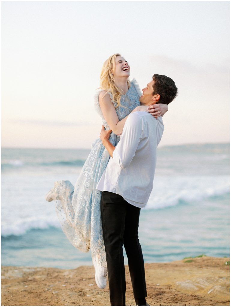 A groom holds his bride in the air as she smiles and laughs in a Christina Sfez light blue wedding gown, in front of a warm ocean view.