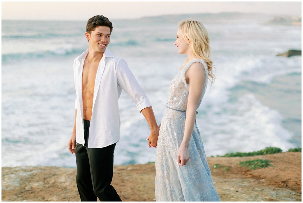 A couple walks along the beach after their elopement by the ocean, holding hands, smiling and looking at each other.