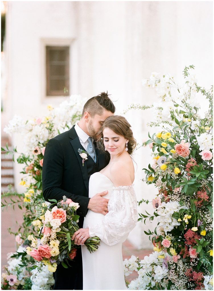 Intimate portrait of married couple outside white building, bride is holding Oak and the Owl coral and yellow floral arrangement, outside Junipero Serra Museum.