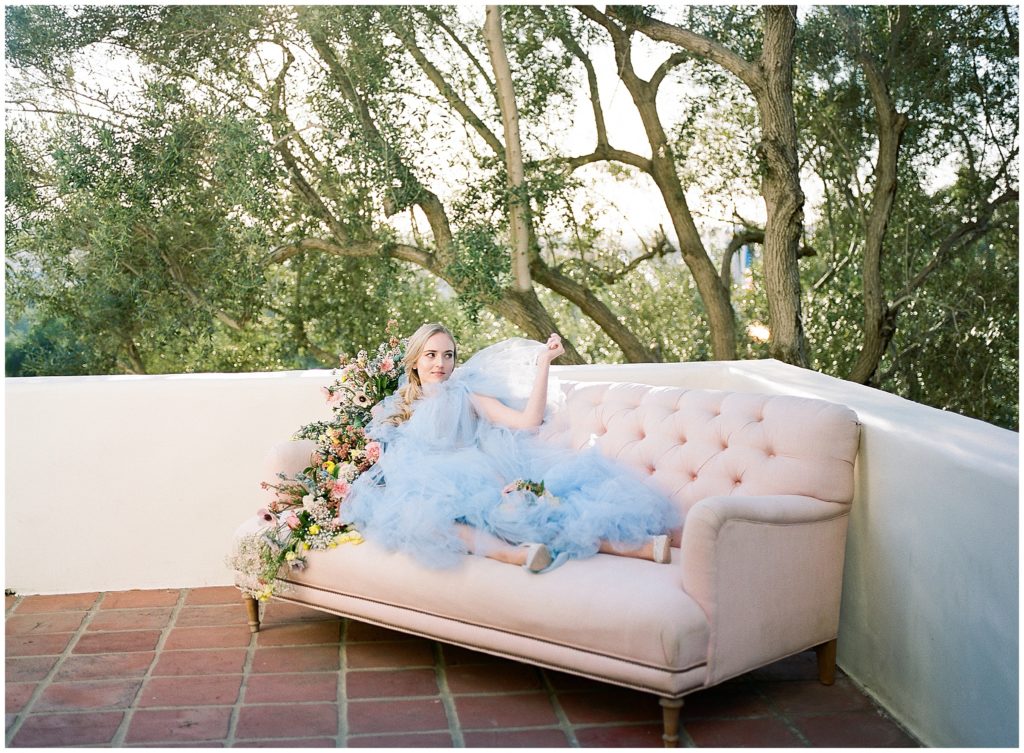 Fine art portrait of bride wearing Elizabeth Dye blue wedding gown, relaxing on pink couch outside on balcony in San Diego