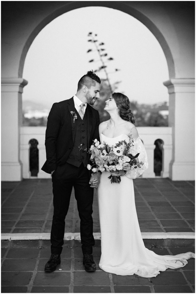 A just married couple smiles at each other after their destination spring wedding, under an archway; the bride is holding a large floral arrangement designed by Oak and the Owl