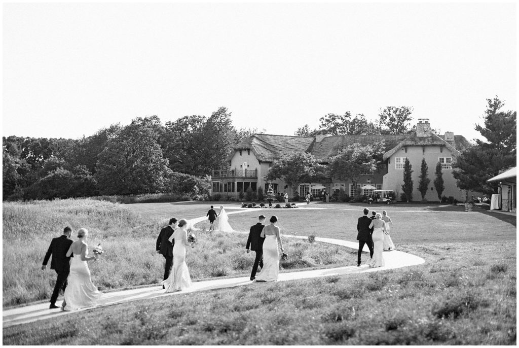Black and white image of bride, groom and wedding party walking back to venue after outdoor wedding ceremony in Minnesota