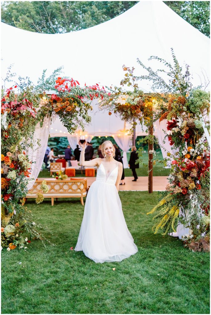 Bride raises a glass of champagne under a floral arch at the entrance of their sailcloth tent at upscale backyard wedding