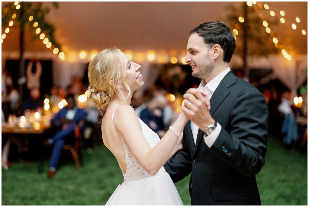 Couple laughs during first dance in tent reception with string lights in the background from their tent reception